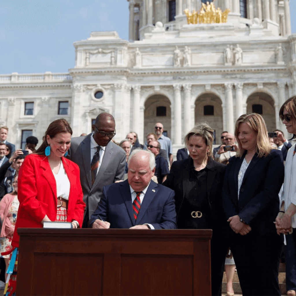 Governor Tim Walz sits in front of the Minnesota State Capitol Building signing a bill into law. He is joined by Lieutenant Governor Peggy Flanagan, Speaker Melissa Hortman, Senate President Bobby Joe Champion, and a crowd in the background.