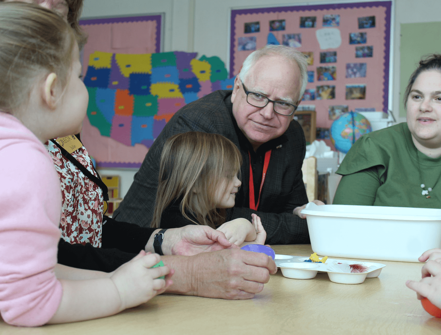 Governor Tim Walz sits in a Minnesota public school classroom next to a young student who is sharing their work with him.