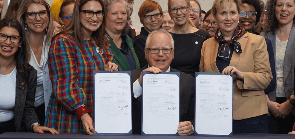 Governor Tim Walz is sitting and holding three recently signed bills in front of him with Lieutenant Governor Flanagan and a crowd of supporters.